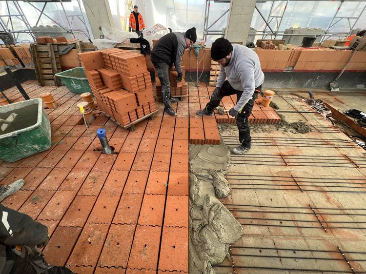 „Facharbeiter verlegen Mauerziegel mit Mörtel auf einer Baustelle in einem gewerblichen Gebäude; im Hintergrund verschiedene Werkzeuge und Baumaterialien, die für die Gebäudetechnik im Bereich Sanitär-, Heizungs- und Klimatechnik verwendet werden.“.