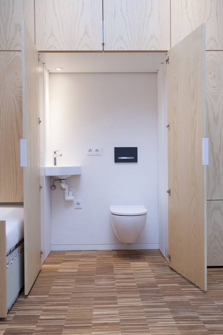 „Modernes, barrierefreies Badezimmer mit wandhängendem WC und kleinem Waschbecken, eingepasst in einen kompakten Raum mit Holzverkleidung auf gefliestem Boden – optimiert für Platzersparnis und einfache Zugänglichkeit in der Gebäudetechnik.“.
