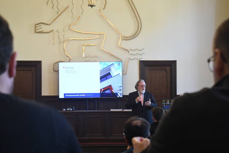 „Referent präsentiert auf einer Fachkonferenz zu Gebäudetechnik, Großbildschirm zeigt Diagramme zu Energieeffizienz, Publikum bestehend aus TGA-Planern und SHK-Fachhandwerkern.