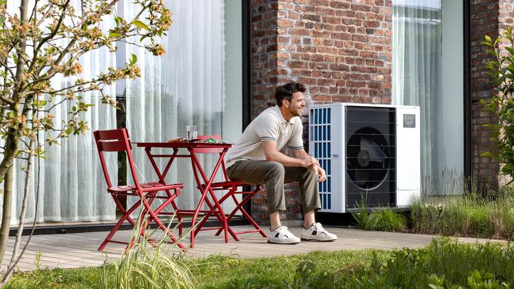 Ein Fachhandwerker inspiziert eine wandmontierte Klimaanlage auf einer Terrasse, umgeben von Backsteinwänden und großen Fenstern, als Teil der energetischen Gebäudeoptimierung.