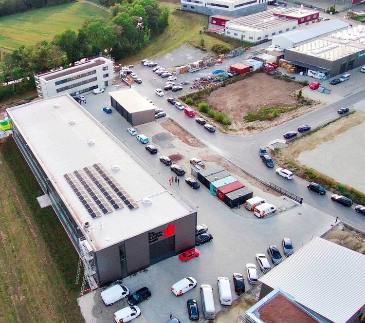 Luftaufnahme eines Gewerbegebiets mit industriellen Gebäuden, die mit Solarpaneelen ausgestattet sind, sowie parkende Fahrzeuge. Umgeben von Grünflächen, zeigt das Bild nachhaltige Energieeffizienzlösungen für die Gebäudetechnikbranche.