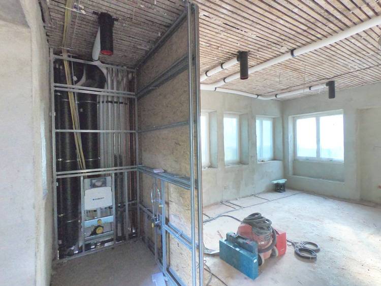 „Baustelle eines Gebäudetechnikprojekts mit offener Verkabelung und Isolierung an den Wänden, verschiedenen Werkzeugen und Geräten auf dem Boden. Große Fenster im Hintergrund lassen natürliches Licht in den Raum strömen, während die Installation von modernen Heizungssystemen und energieeffizienter Klimatechnik vorbereitet wird.