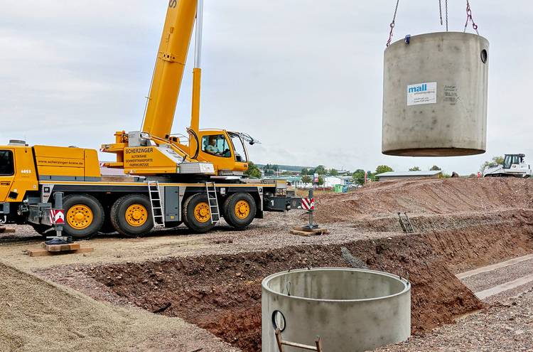 Ein Kran hebt einen Betonabschnitt an einer Baustelle, umrahmt von Kies- und Erdboden. Im Hintergrund sind Baugerüste und Gebäudekomponenten für energieeffiziente Heizungs- und Belüftungssysteme sichtbar, die wichtige Lösungen für Entscheider in der Gebäudetechnik darstellen.