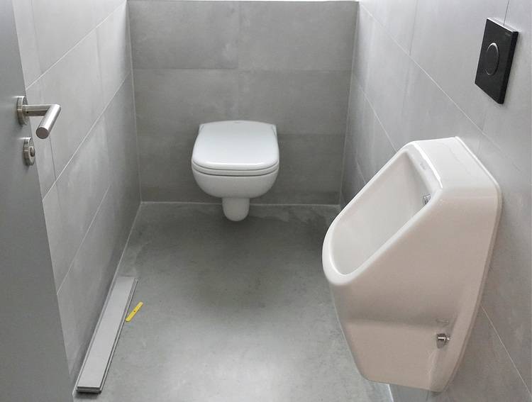 „Barrierefreies WC mit wandhängendem WC und Urinal, graue Fliesen, ausgestattet für die Bedürfnisse moderner Gebäudetechnik und Sanitärlösungen.
