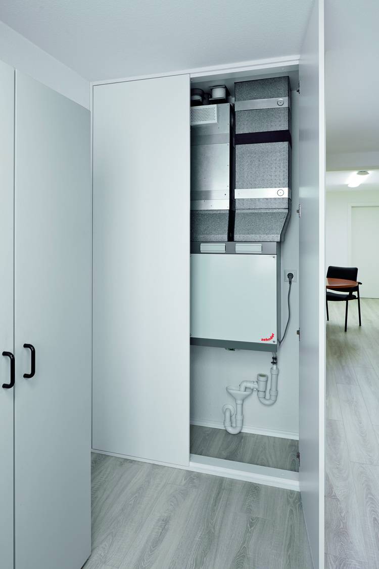 Beschreibung eines kleinen Technikraums mit installierter Lüftungseinheit an der Wand und sichtbaren Rohrleitungen am Boden, fokussiert auf effiziente Gebäudetechniklösungen für Fachkräfte und Entscheider in der Planung und Umsetzung von Energieeffizienzmaßnahmen.