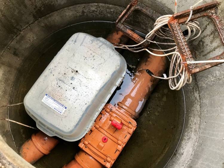 Technische Detailaufnahme eines unterirdischen Versorgungsschachts mit verschiedenen Versorgungsleitungen und einem teilweise im Wasser stehenden Kunststoffbehälter, relevant für Entscheider in der Gebäudetechnik zur Optimierung von Energieeffizienz und Wartungsstrategien.