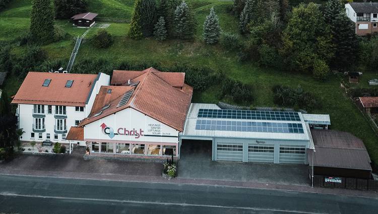 Natürlich, hier ist ein optimierter ALT-Text für das beschriebene Bild: „Vogelperspektive auf ein modernes Gebäude mit roten Dächern, rechts integrierte Solarpaneele zur nachhaltigen Energieerzeugung, umgeben von grüner Landschaft. Relevantes Beispiel für energieeffiziente Bauweise in der Gebäudetechnikbranche.