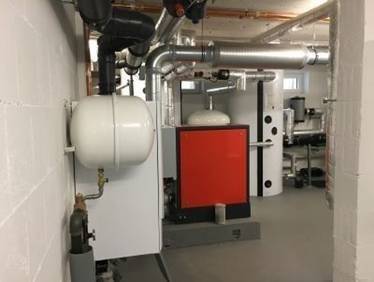 „Kellerraum mit moderner Heizungsanlage, bestehend aus einem energieeffizienten, roten Wärmekessel, einem weißen Pufferspeicher und verzweigten Metallrohrleitungen für optimierte Energienutzung und -verteilung.