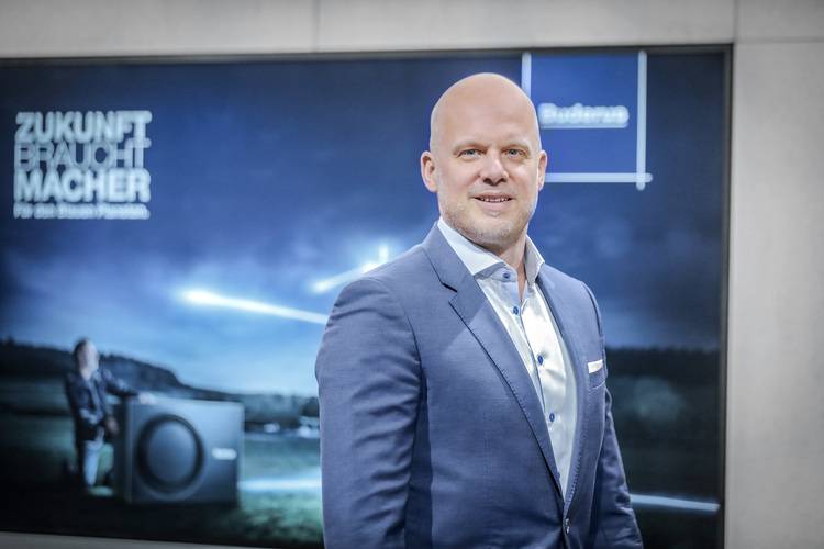 „Fachmann im blauen Anzug, der sich vor einem modernen Hintergrund präsentiert; im Bild befinden sich deutsche Schlagworte zur Gebäudetechnik wie Energieeffizienz, Klimasysteme und erneuerbare Energien.