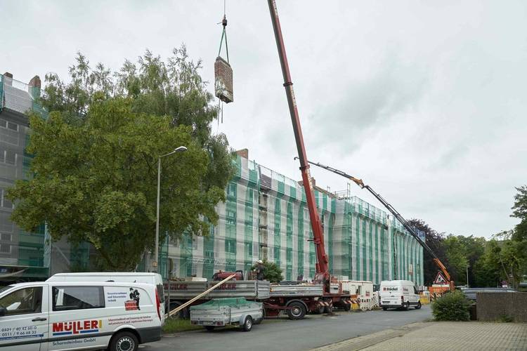 „Baustelle mit Kränen, die Baumaterialien zu einem mit Gerüsten und grünem Netz eingerüsteten Gebäude heben, symbolisiert fortschrittliche Gebäudetechnik und Energieeffizienzmaßnahmen in der Bauausführung.“.