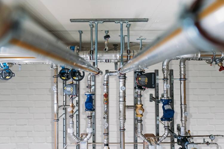 Optimierter ALT-Text: „Vernetzte Rohrleitungen und Absperrventile eines modernen Heizungs- und Sanitärsystems, fachgerecht auf einer weißen Wand in einem Technikraum installiert – Ansicht aus der Perspektive eines Gebäudetechnikers.“.