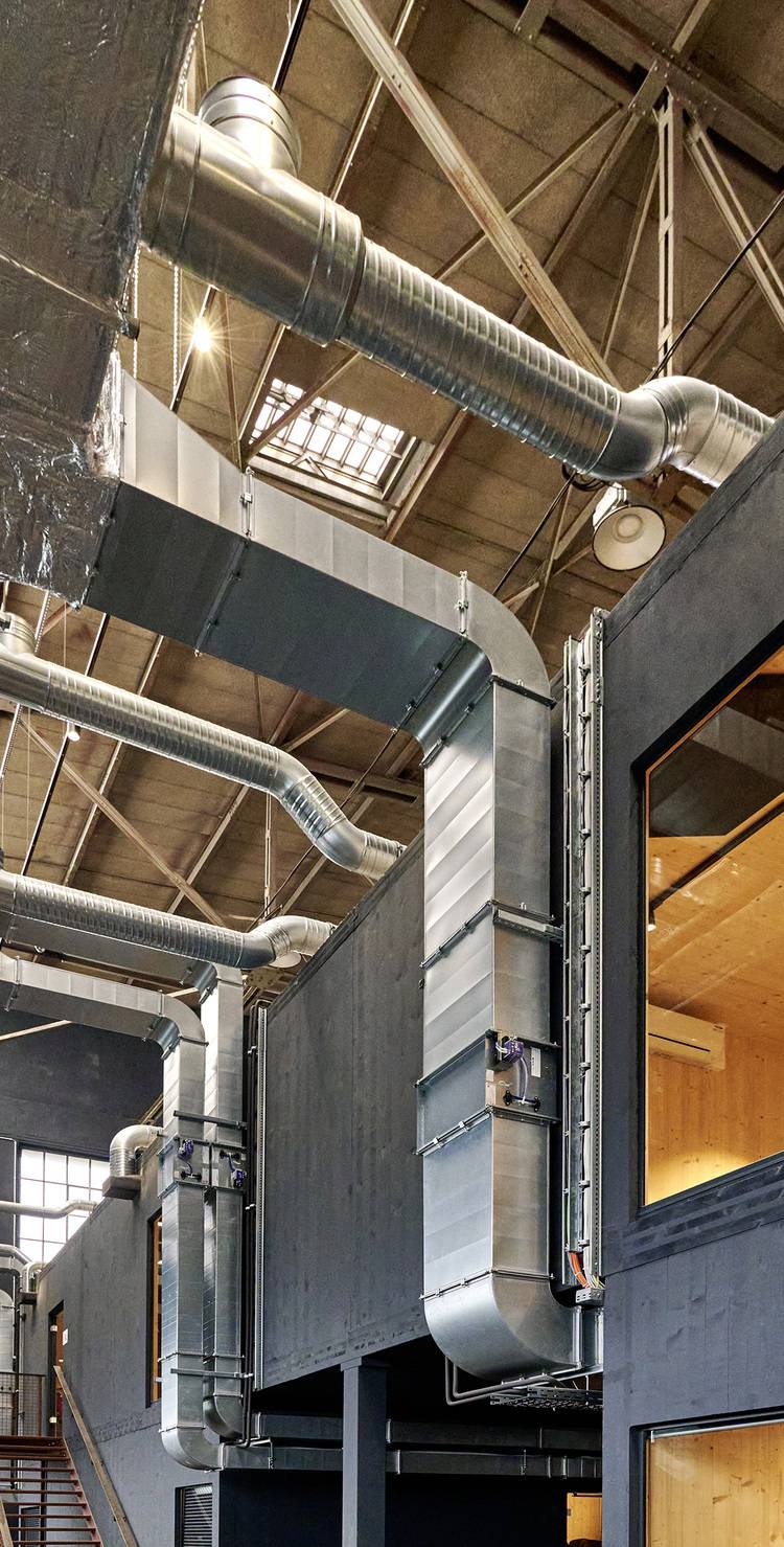 Moderne Innenansicht eines Industriegebäudes mit sichtbarer Metallrohrführung und hoher Decke, ausgestattet mit Oberlichtern zur natürlichen Belichtung. Die schwarze Wandgestaltung unterstreicht das energieeffiziente Design und die Nutzung erneuerbarer Energien in der Gebäudetechnik.
