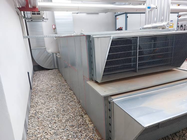 Optimierter ALT-Text: „Lüftungskanäle aus Metall mit robusten Gittern auf Kiesboden in einem hellen Technikraum eines Gewerbegebäudes, beispielhaft für moderne industrielle Klimatechnik und Gebäudetechnik-Anwendungen.