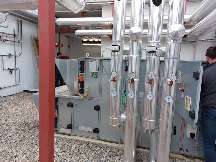 Optimierter ALT-Text: „Groß dimensionierte Heizungs-, Lüftungs- und Klimaanlage (HLK) mit verzweigten Metallrohren und Messinstrumenten, zwei Gebäudetechniker prüfen Anlagenkomponenten in einem Technikraum mit Kiesboden – typisches Arbeitsumfeld für TGA-Fachkräfte.“.