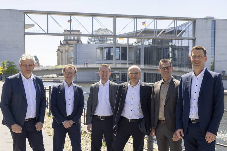 Alt-Text-Vorschlag: „Sechs Männer im Anzug diskutieren im Freien am Flussufer; im Hintergrund moderne Bürogebäude und Brücken – Netzwerktreffen von Entscheidern der Gebäudetechnik-Branche bei einer Fachveranstaltung.“.