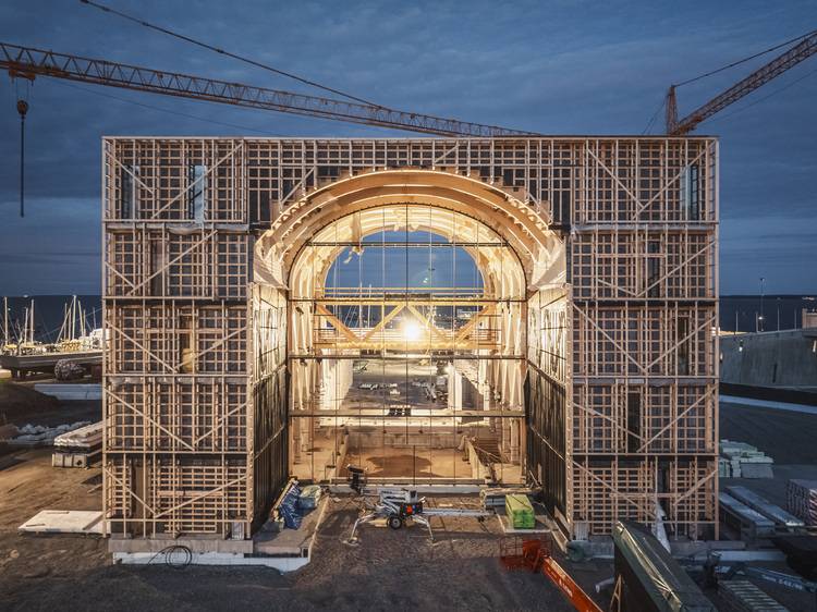 Bisheriger Alt-Text: „Ein großes, beleuchtetes Gebäude im Bau mit Holzgerüsten und Kränen in der Abenddämmerung.“ Optimierter ALT-Text für haustec.de (zielgruppen- und themenrelevant): „Beleuchtete Großbaustelle eines Bürogebäudes im Rohbau mit Holzgerüst und zwei Kränen bei Dämmerung; Fokus auf innovative Gebäudetechnik, Energieeffizienz und moderne TGA-Lösungen im Neubau.