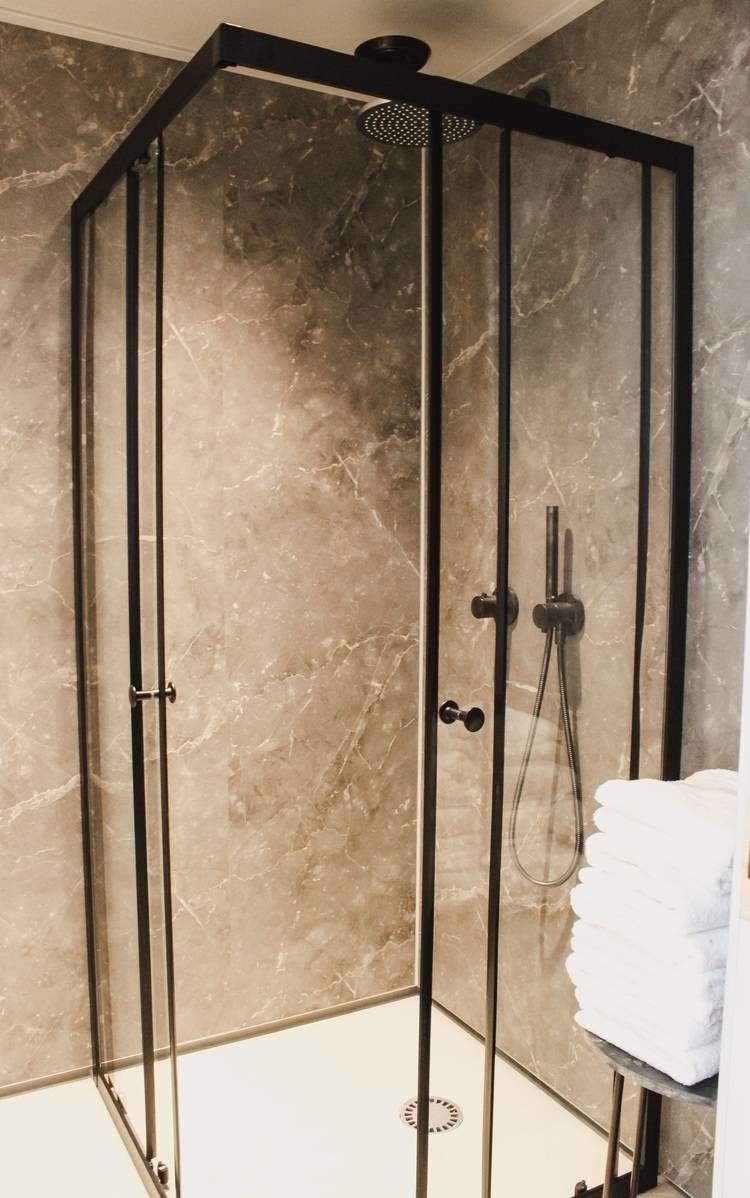 Optimierter, themenrelevanter ALT-Text: Barrierefreie, moderne Duschkabine mit bodengleichem Einstieg und schwarzem Rahmen vor grauen Feinsteinzeugwänden in einem energieeffizienten Badezimmer; daneben gestapelte, weiße Handtücher als Ausstattungselement einer zeitgemäßen Sanitärlösung für Wohn- und Gewerbeimmobilien. Begründung: Der Text legt den Fokus auf Barrierefreiheit (zentrales Thema der Gebäudetechnikbranche), benennt die Materialität (Feinsteinzeug statt Marmor/Marble für den Branchentonus), erwähnt die Energieeffizienz als aktuellen Trend sowie Anwendungsbereich Wohn- und Gewerbeimmobilien – alles relevante Schlüsselbegriffe für Ihre Zielgruppe aus Entscheider:innen der Gebäudetechnik.