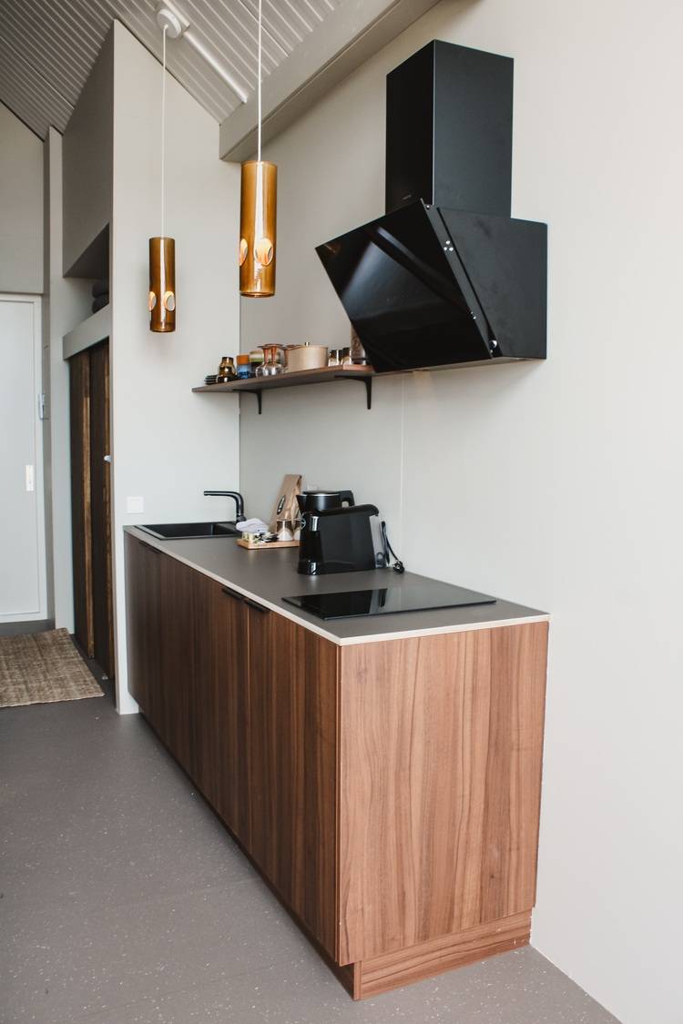 Optimierter ALT-Text: "Moderne Einbauküche mit energieeffizienten Geräten und eingebauter kontrollierter Wohnraumlüftung, offenem Holzregal, zwei LED-Pendelleuchten über der Kücheninsel; Beispiel für nachhaltige Gebäudetechnik und intelligente Lichtplanung im Wohnbereich.