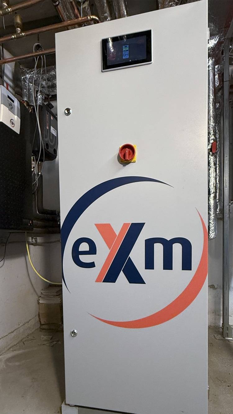 Optimierter ALT-Text: Weißer Industrie-Schaltschrank mit integriertem Touchscreen, rotem Not-Aus-Schalter und „exm“-Beschriftung in einem Technikraum, eingesetzt zur Steuerung und Überwachung gebäudetechnischer Anlagen.