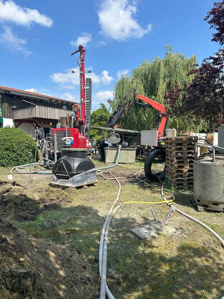 ALT-Text-Vorschlag: „Bauarbeiter installieren Erdwärmesonden auf einer Baustelle für eine Gebäudetechnik-Anlage; zu sehen sind Bohrgeräte, Rohrleitungen und Fachkräfte bei der Vorbereitung eines energieeffizienten Versorgungssystems neben Bestandsgebäuden und Bäumen.