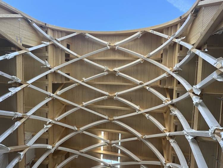 Natürlich! Hier ein optimierter, themenrelevanter Alternativtext für das beschriebene Bild für haustec.de: ALT-Text: „Gebogenes Dach im modernen Gebäudebau mit tragender Holzbalkenkonstruktion in Kreuzstruktur; Fokus auf nachhaltige Baustoffe und innovative Tragwerksplanung für energieeffiziente Gebäudehüllen unter freiem Himmel.“ Dieser Alt-Text ist spezifisch und branchenrelevant formuliert und spricht die Themen Nachhaltigkeit, Tragwerksplanung und Gebäudehülle an – Bereiche, die Entscheider der Gebäudetechnik interessieren.