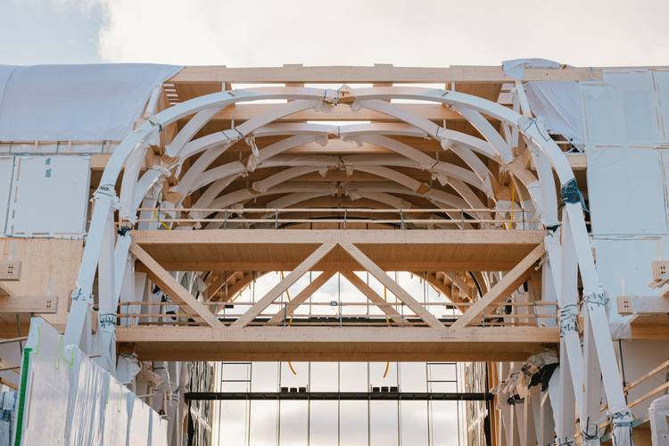 Gerne, hier ist ein optimierter, themenrelevanter ALT-Text für das beschriebene Bild im Kontext der Gebäudetechnik-Branche: Alt-Text: Holzbalken und gebogene Holzträger bilden das Tragwerk eines energieeffizienten Gebäudes im Rohbau; im Hintergrund Himmel mit vereinzelten Wolken.