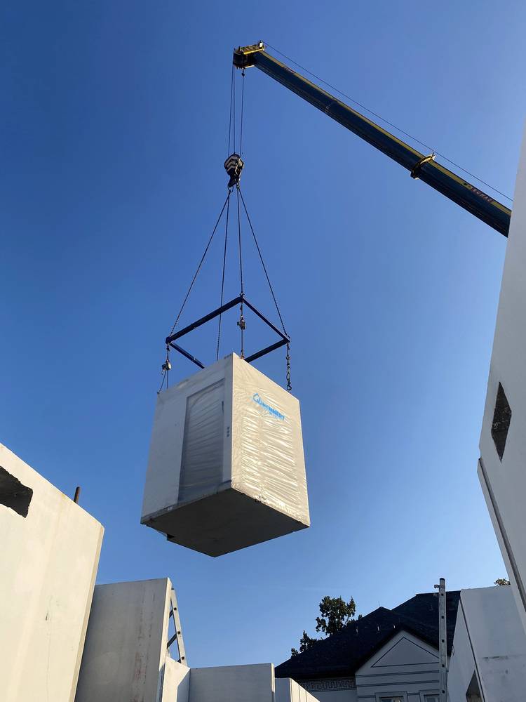 Alt-Text-Vorschlag: "Ein Kran hebt einen großen, weiß verpackten Lüftungs- oder Heiztechnikcontainer über eine Baustelle für ein neues Energieeffizienzgebäude; im Hintergrund arbeiten Haustechnik-Fachkräfte an der Installation moderner Gebäudetechnik unter freiem Himmel.
