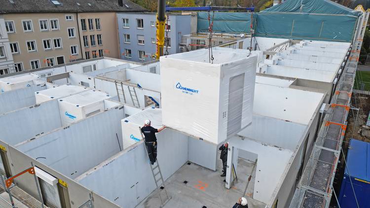 Optimierter ALT-Text: "Ein Kran hebt ein vorgefertigtes Technikmodul auf die Dachfläche eines Gebäudes in der Rohbauphase, während Installateure das Modul für den Anschluss an die haustechnischen Anlagen positionieren." Begründung: Dieser Alt-Text beschreibt den Vorgang spezifisch im Kontext der Gebäudetechnik und lenkt die Aufmerksamkeit auf modulare Installation und Integration von Techniksystemen - zentrale Themen für die Zielgruppe von haustec.de.