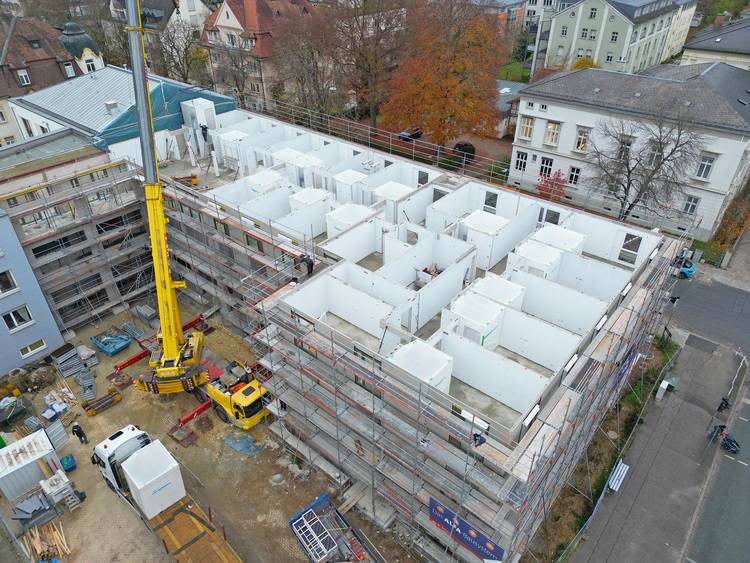 ALT-Text-Vorschlag (optimiert für haustec.de und die genannte Zielgruppe): "Luftaufnahme eines Bürogebäudes im Rohbau mit Kran und umfassendem Gerüst; sichtbar sind die Installation von Heizungs-, Lüftungs- und Sanitäranlagen durch Fachkräfte, Fokus auf Gebäudetechnik während der Bauphase.