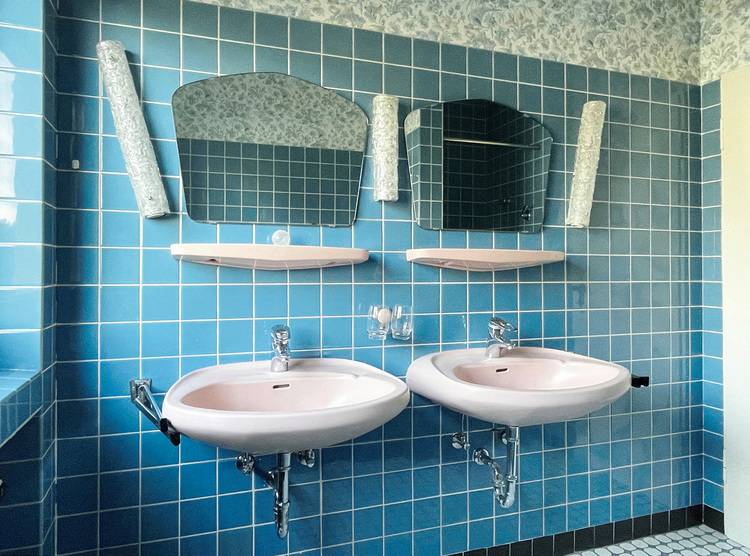 Gerne, hier ein optimierter ALT-Text mit Bezug zu Gebäudetechnik und der fachlichen Zielgruppe: Zwei wandhängende Waschtische mit berührungslosen Armaturen und Spiegeln vor blau gefliester Badezimmerwand; Fokus auf moderne Sanitärtechnik und barrierefreie Badgestaltung für öffentliche oder gewerbliche Gebäude.