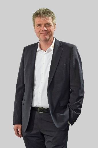 Neuer, optimierter ALT-Text: "Geschäftsmann im dunklen Anzug und weißem Hemd, Vertreter eines TGA-Unternehmens, steht lächelnd vor neutralem Hintergrund - symbolisiert Entscheider in der Gebäudetechnikbranche.