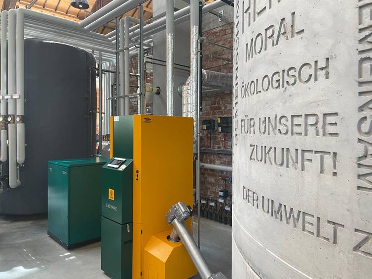 Optimierter ALT-Text: Technikraum mit modernen Rohrleitungen, wandhängendem Kessel in Gelb-Grün für Heizung und Warmwasser sowie Betonwand mit deutscher Beschriftung zu Energieeffizienz und Umweltschutz in der Gebäudetechnik.