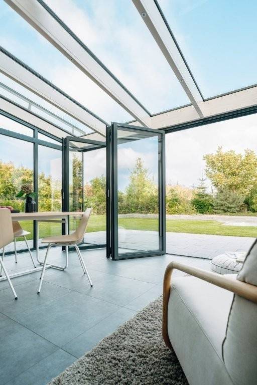 Barrierefrei optimierter ALT-Text für haustec.de (B2B Zielgruppe, Gebäudetechnik): "Wintergarten mit vollflächiger Verglasung und energieeffizienter Gebäudehülle, geöffnete Türen zur Gartenanlage; im Innenraum funktionale Einrichtung mit Tisch, Stühlen und Sofa - Beispiel moderner Wohnraumerweiterung unter Berücksichtigung energetischer und gebäudetechnischer Anforderungen.