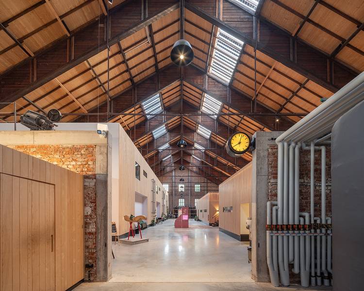 Alt-Text-Vorschlag für haustec.de (optimiert für die Zielgruppe und den Kontext Gebäudetechnik): "Modernisiertes Industriegebäude mit freiliegenden Heizungsrohren, großformatiger Uhr, offenem Holzdachstuhl sowie sichtbaren Backsteinwänden - Beispiel für energieeffiziente Sanierung und zeitgemäße Gebäudetechnik.