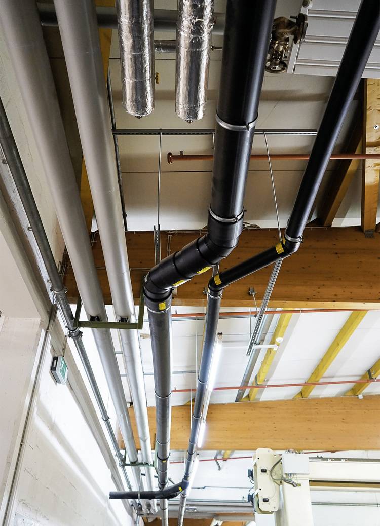 Installationsbeispiel: Schwarz-silberne Rohrleitungen für Heizungs- oder Klimatechnik verlaufen unter Holzdecken mit Metallträgern in Industriehalle.