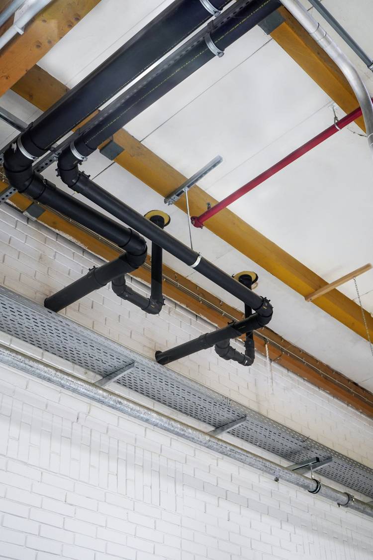 Moderne Gebäudetechnik: Schwarze Rohrleitungen und Metallkanäle verlaufen auf weißer Ziegeldecke mit Holzbalken und rotem Installationskanal.