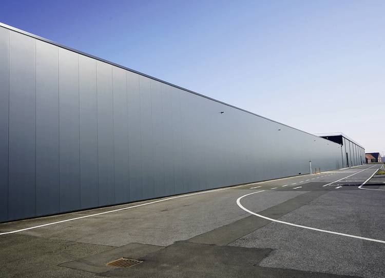 Moderne, lang gestreckte Industriehalle als Lagergebäude für Gebäudetechnik-Gewerke, vor leerer Zufahrtsstraße und klarer Himmelkulisse.