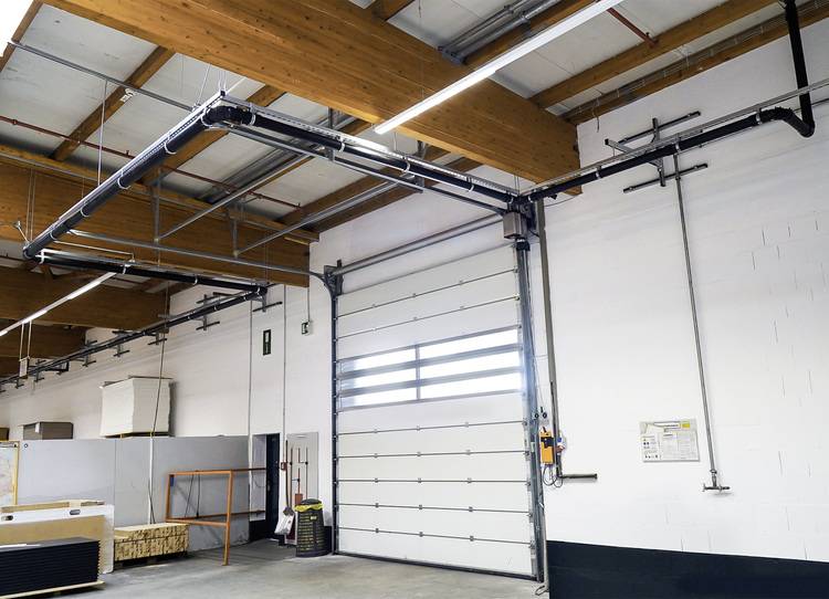 Innenansicht eines Lagers mit geschlossener Industrietoranlage, sichtbaren Dachträgern sowie gelagerten Kisten und haustechnischem Equipment.