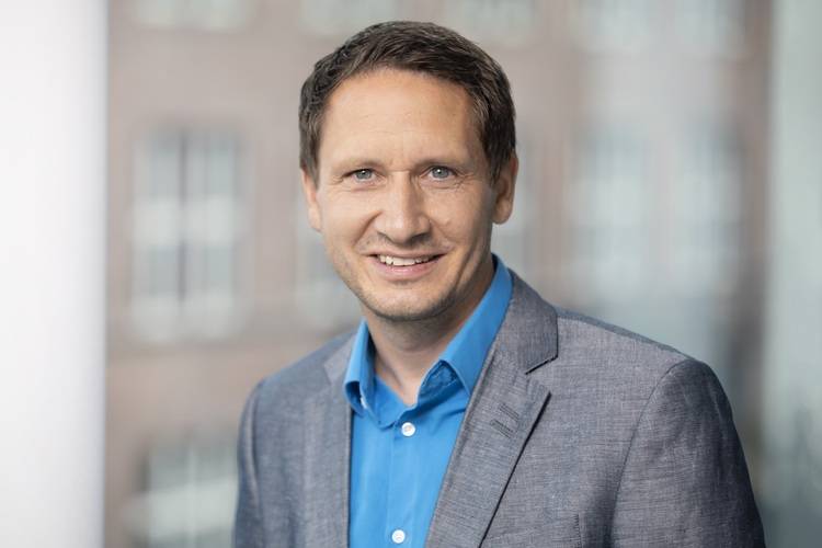 Fachplaner im grauen Sakko und blauem Hemd bei einer Branchenveranstaltung für Gebäudetechnik, unscharfe Fenster im Hintergrund.