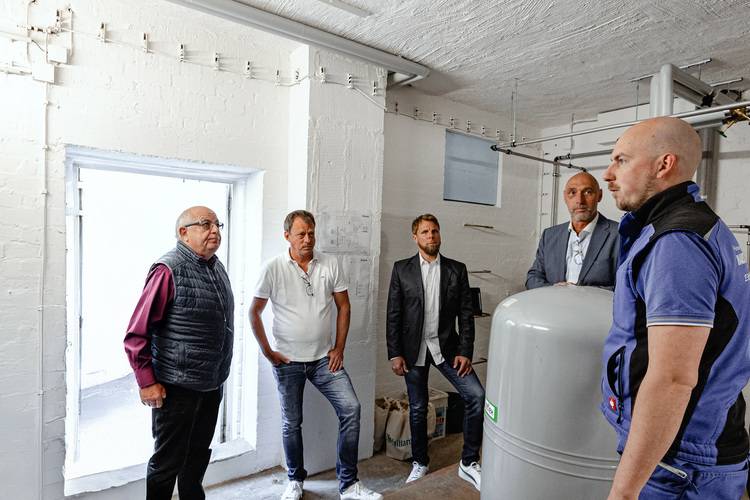 Fünf Fachleute diskutieren in einem hellen Technikraum vor Industrieanlagen und Rohrleitungen über gebäudetechnische Systeme.