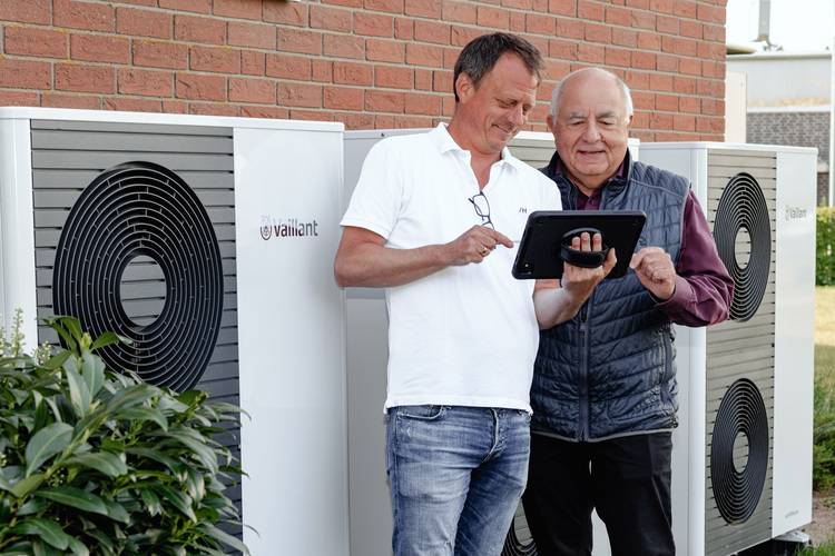 Zwei Fachkräfte prüfen gemeinsam über ein Tablet moderne Wärmepumpenanlagen vor einem Ziegelgebäude im Kontext Gebäudetechnik-Effizienz.