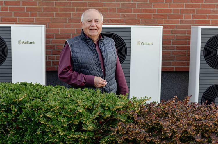 Fachmann der Gebäudetechnik präsentiert moderne Wärmepumpen vor Ziegelwand, Fokus auf innovative Heiztechnik im Außenbereich.