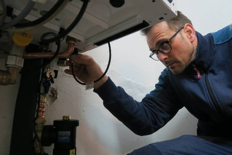 Fachhandwerker in blauer Arbeitskleidung prüft Rohrleitungen unter Heizkessel, Inspektion und Wartung im Bereich Gebäudetechnik.