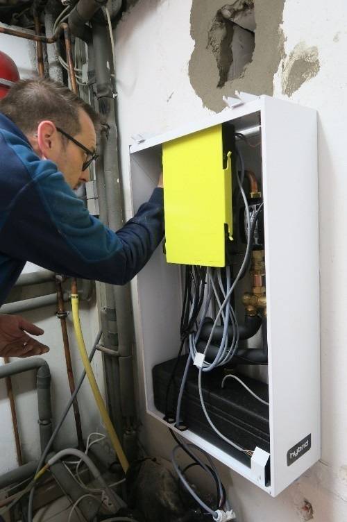 Fachkraft prüft wandmontierten Installationskasten mit freiliegenden Rohrleitungen und Kabeln, Fokus auf Gebäudetechnik-Installation.