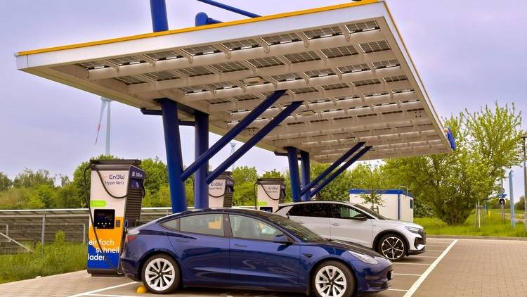 Mehrere E-Autos laden an solarbetriebenen Ladestationen mit großem Überdach auf Parkplatz, umgeben von Begrünung, Fokus auf Energieeffizienz.