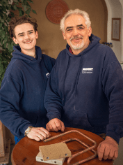 Zwei Fachkräfte in blauen Hoodies präsentieren an einem Holztisch verschiedene Metallbauteile für die Gebäudetechnikbranche.