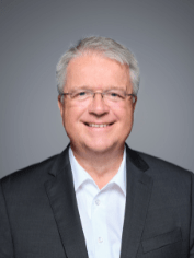 Porträt eines älteren Fachmanns der Gebäudetechnikbranche mit Brille, grauen Haaren und dunklem Sakko vor neutralem Hintergrund.