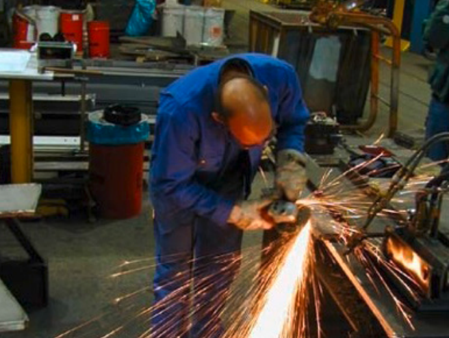 Fachkraft in blauer Arbeitskleidung führt mit Winkelschleifer Metallbearbeitung in Werkstatt durch, helle Funken fliegen - Fokus auf Gebäudetechnik-Handwerk.