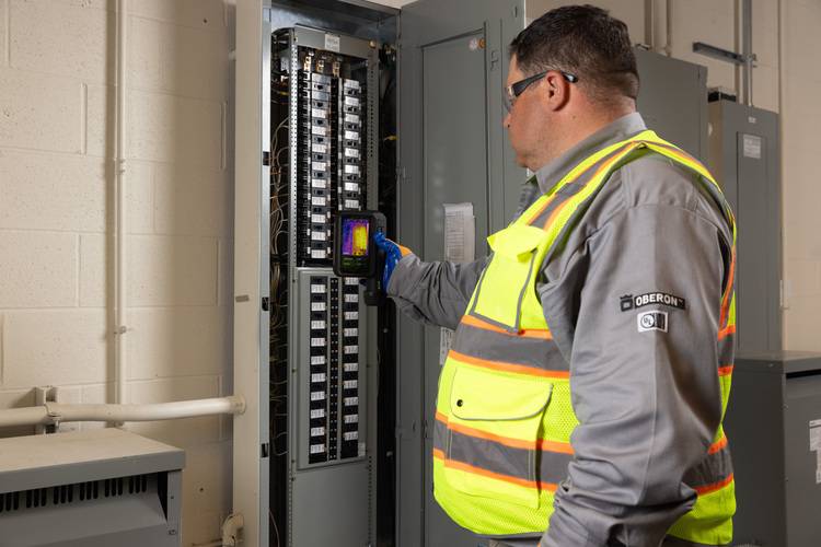 Fachkraft prüft mit mobilem Prüfgerät einen geöffneten Elektroschaltkasten in einem Technikraum zur Sicherstellung normgerechter Gebäudetechnik.