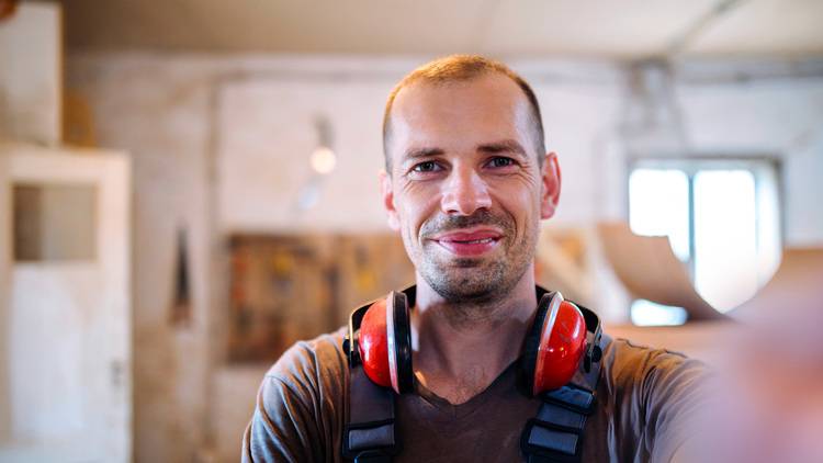 Facharbeiter mit kurzem Haar und roten Gehörschutz-Kapseln um den Hals steht in einer Gebäudetechnik-Umgebung, geeignet für SHK-Berufe.